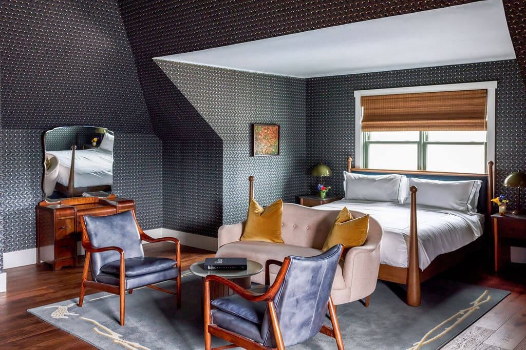 4 New Nashville Boutique Hotels That Should Be on Your Radar - 3 A stylish bedroom with dark patterned wallpaper, a wooden bed with white bedding, a tan loveseat, two blue armchairs, a small round table, and a dressing table with mirror.