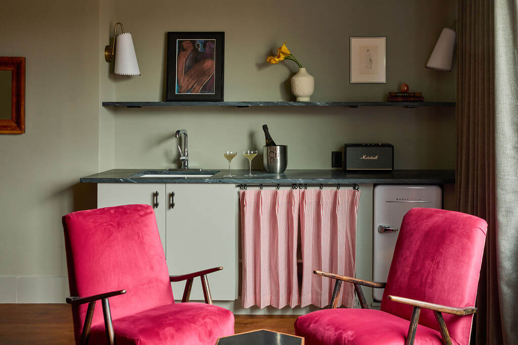 4 New Nashville Boutique Hotels That Should Be on Your Radar - 4 Small kitchenette with a sink, two pink armchairs, a fridge, champagne glasses, and art on the shelves and wall. Pink striped curtain covers the lower cabinet.