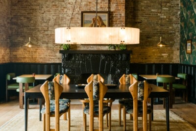 A dining room with a long wooden table, eight chairs, a large hanging light fixture, exposed brick wall, and a black fireplace with a framed portrait above it.