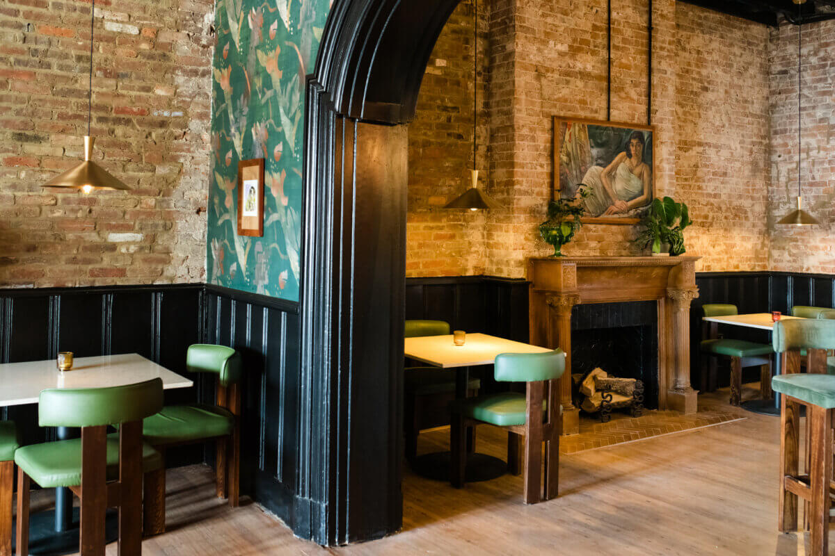 4 New Nashville Boutique Hotels That Should Be on Your Radar - 9 A cozy restaurant interior features green cushioned chairs, white tables, exposed brick walls, a fireplace with a painting above it, and pendant lights.