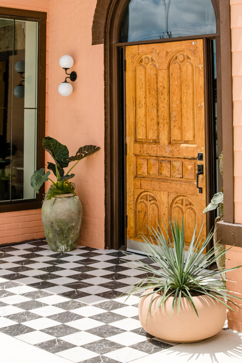 4 New Nashville Boutique Hotels That Should Be on Your Radar - 7 Wooden front door with arched window, flanked by large potted plants on a black-and-white checkered tile floor, beside a peach-colored brick wall.