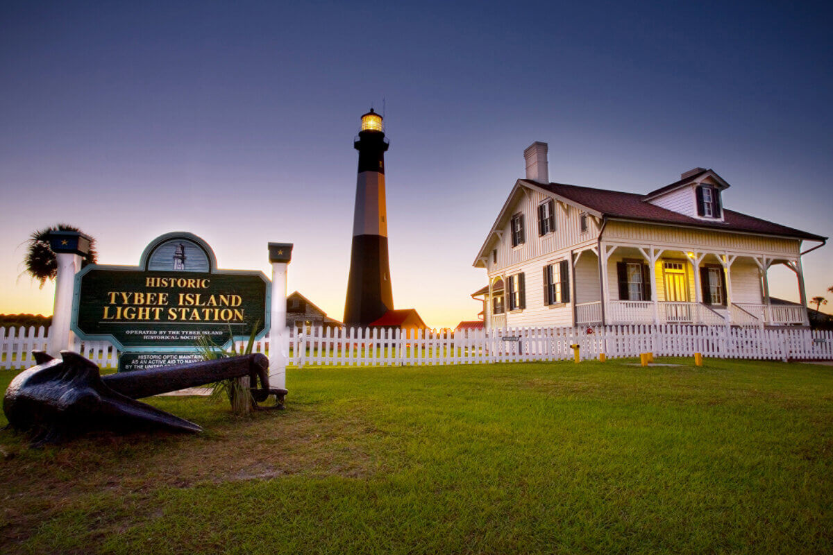 The Best Summer Weekend Getaways in the South - 2 Tybee Island Light Station at sunset, with a white lighthouse, adjacent historic keeper's house, and a sign reading "Tybee Island Light Station" on a grassy lawn—a South favorite for summer weekend trips.