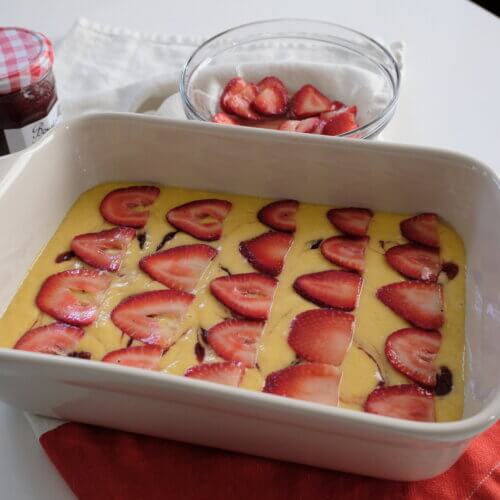 RECIPE: Strawberry Snacking Cake - 3 A baking dish filled with a yellow batter topped with sliced strawberries, next to a bowl of more strawberries and a jar of jam on a white and red cloth.