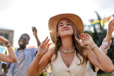 A woman wearing a hat smiles and raises her hands while dancing outdoors, radiating joy with her friends in the background—her glowing look a testament to flawless summer makeup tips.