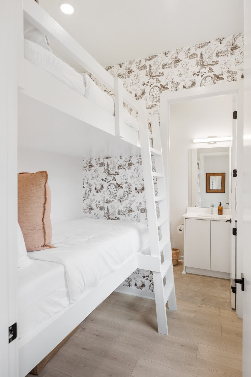 A small room at 1233 Lischey Ave, Nashville, with white bunk beds and a ladder, features patterned wallpaper and a view into a bathroom with a sink and mirror.