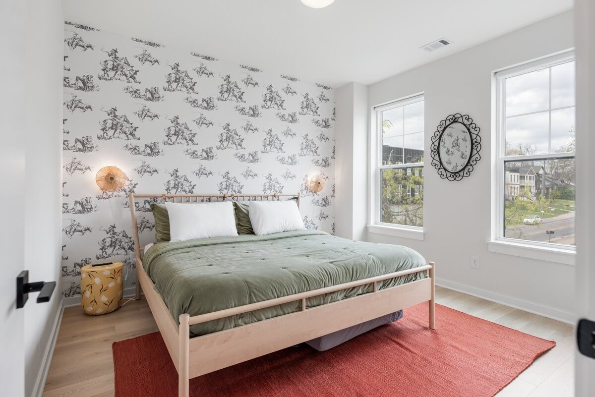 A bright bedroom at 1233 Lischey Ave, Nashville features a wood bed frame, green bedding, two white pillows, a patterned accent wall, wall sconces, two windows, a decorative mirror, and a yellow side table.