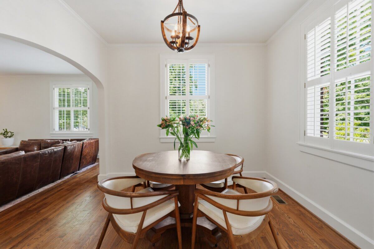 A small dining area with a round wooden table, four chairs, a vase of flowers, and large windows with white shutters welcomes you at 3008 New Natchez Trace in Nashville. A living room with leather sofas is visible through an arched doorway.