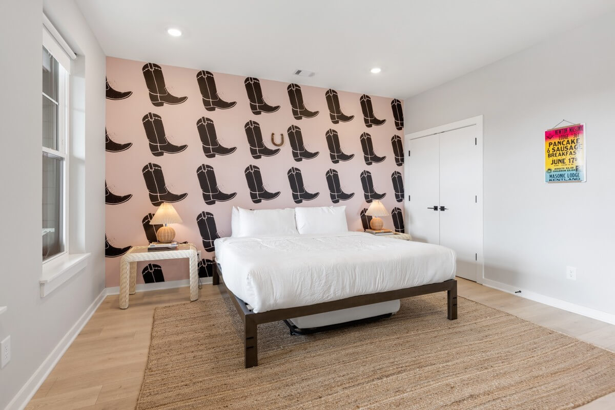A modern bedroom in Nashville TN with a large bed, white bedding, two nightstands with lamps, a pink accent wall featuring black cowboy boot decals, and a colorful poster on a white wall at 1233 Lischey Ave.