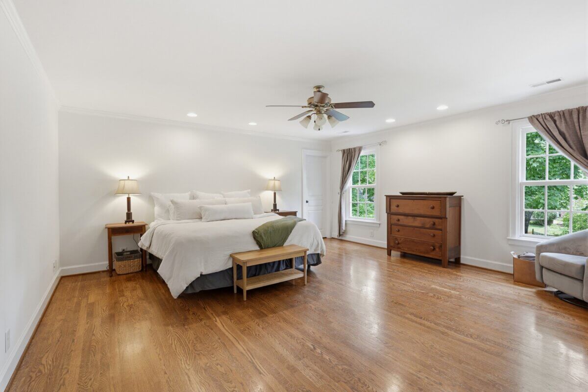 Spacious bedroom in Nashville TN with hardwood floors, a large bed, wooden furniture, ceiling fan, and two windows with brown curtains letting in natural light.