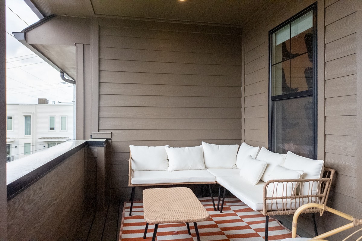 A small covered balcony at 1233 Lischey Ave, Nashville, features a wicker outdoor sectional with white cushions, a wicker coffee table, and a red-and-white striped rug.