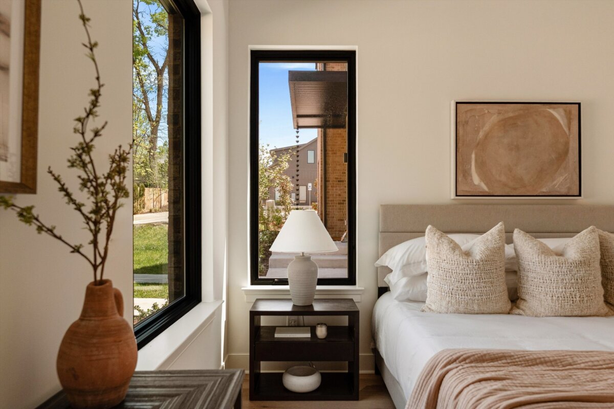 A bedroom at 627 Richmond Bnd, Nashville, features a beige bed, three textured pillows, a lamp on a nightstand, large windows, neutral wall art, and a ceramic vase with branches.
