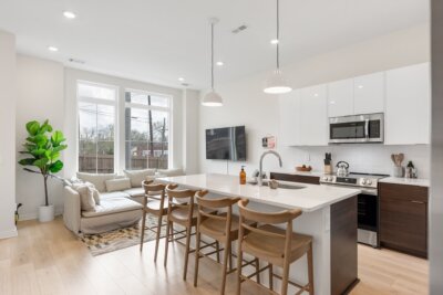 Modern open-concept kitchen and living area at 1233 Lischey Ave, Nashville TN, features white cabinets, island seating, stainless steel appliances, a sectional sofa, large windows, and a potted plant.