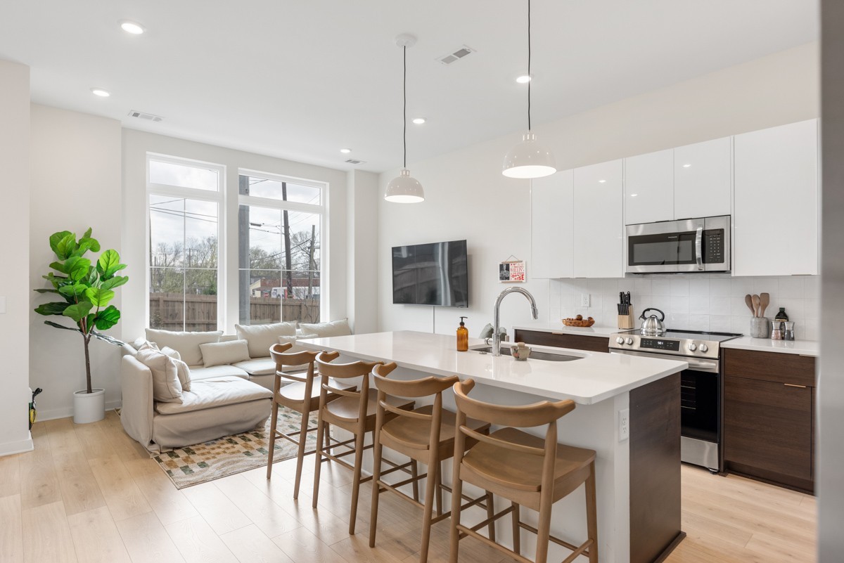 Modern open-concept kitchen and living area at 1233 Lischey Ave, Nashville TN, features white cabinets, island seating, stainless steel appliances, a sectional sofa, large windows, and a potted plant.