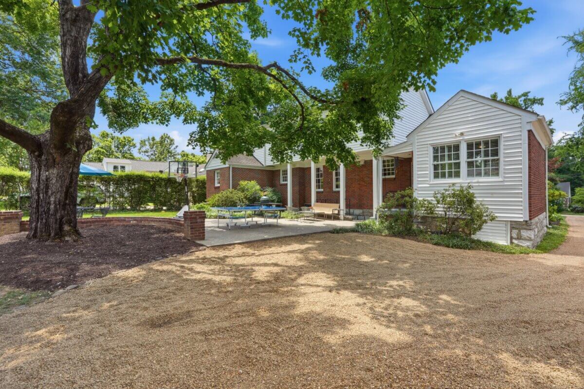 Charming single-story brick house at 3008 New Natchez Trce, Nashville TN, featuring white siding accents, a large tree in the yard, gravel driveway, outdoor seating, and lush landscaping—perfect for real estate seekers under a clear sky.