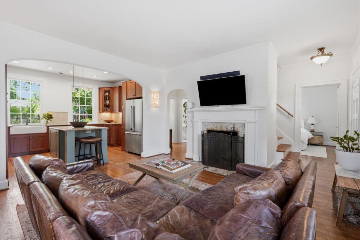 Open-concept living room in New Natchez Trce, Nashville TN, with brown leather sofas, a coffee table, fireplace with TV above, hardwood floors, and a view into the kitchen featuring stainless steel appliances.