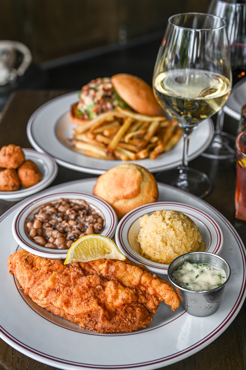A plate with fried fish, hush puppies, beans, cornbread, rice, a lemon wedge, and tartar sauce—classic Nashville Lunch comfort food. In the background, a burger with fries and a glass of white wine are visible.