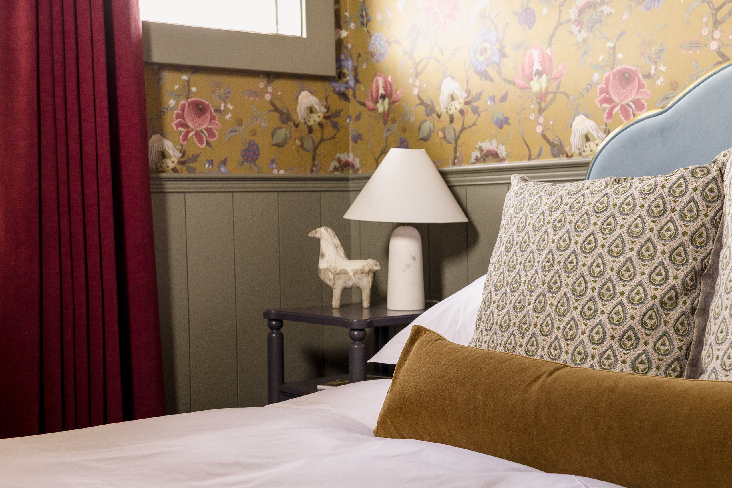 A neatly made bed with patterned pillows, a brown cushion, a lamp, and a decorative horse figurine sits beside floral wallpaper and red curtains in this charming Sylvan Park boarding house.