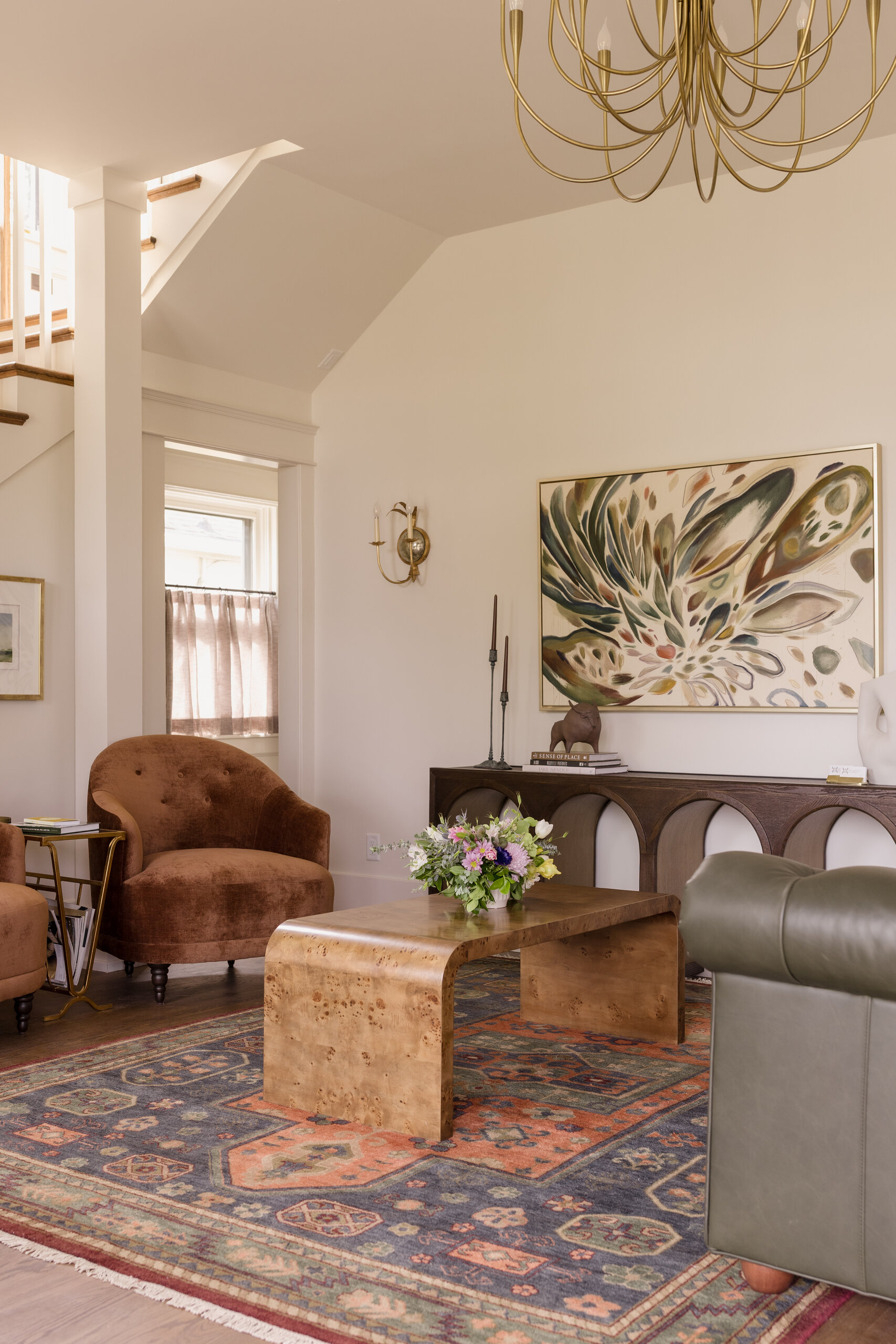 A living room at the Sylvan Park Boarding House features a patterned rug, wood coffee table, velvet chairs, green sofa, wall art, and a gold chandelier illuminated by natural light.
