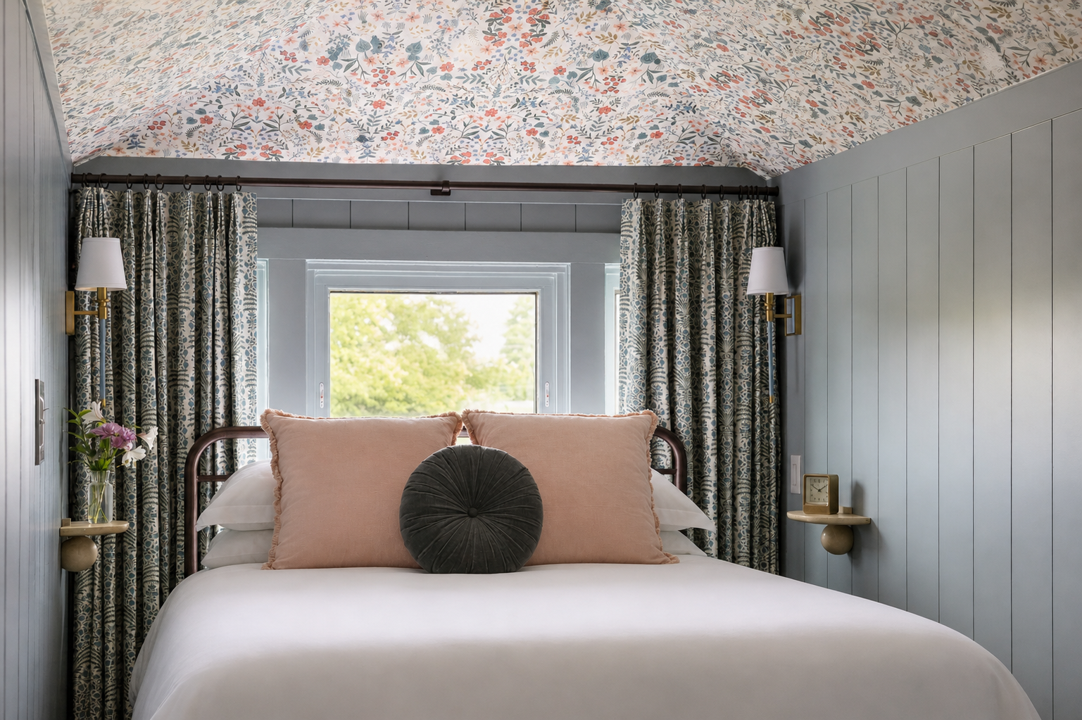 A neatly made bed with peach and dark gray cushions sits under a floral-patterned sloped ceiling at the Sylvan Park Boarding House, flanked by wall sconces and draped curtains, with a window at the head.
