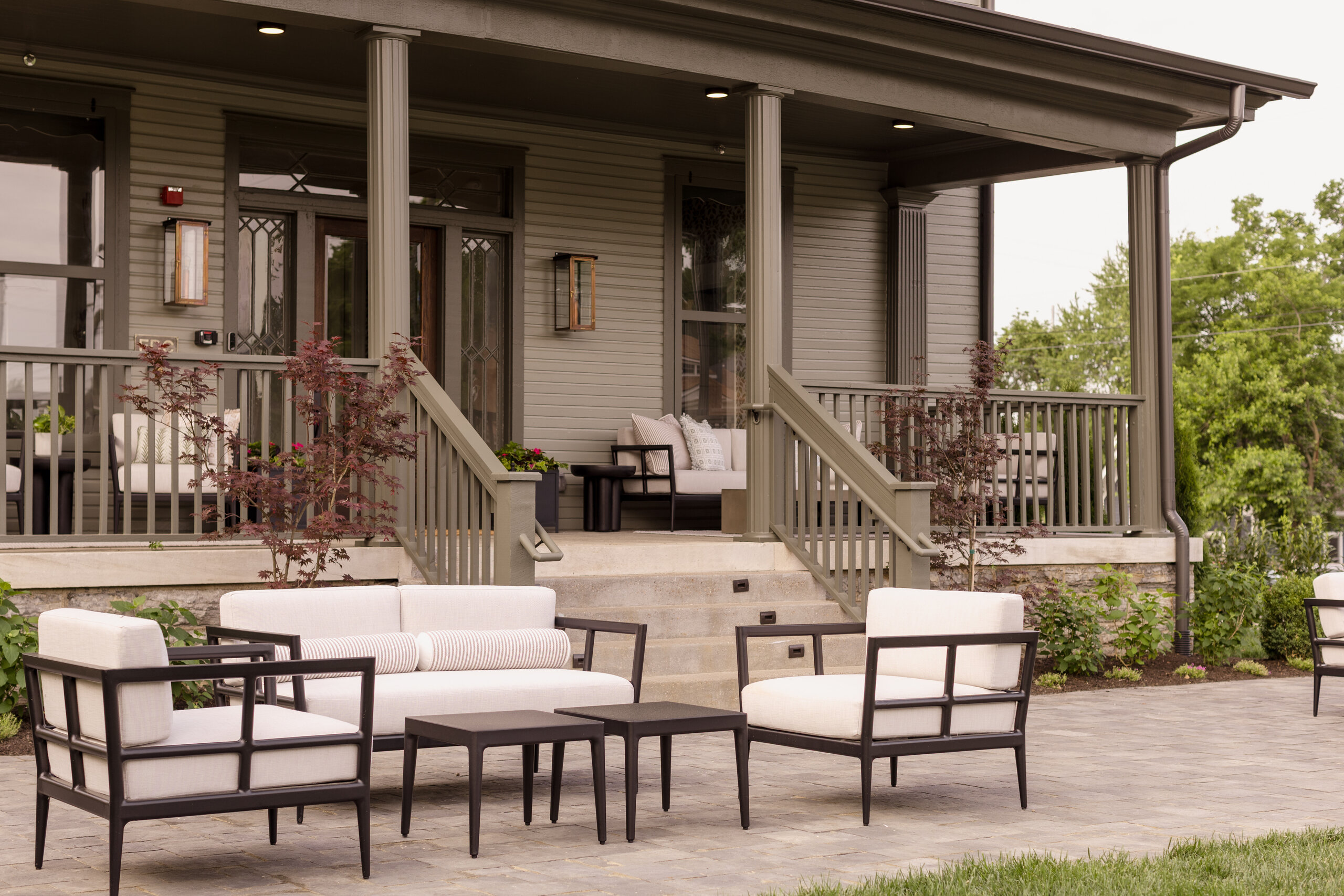 Patio with cushioned outdoor furniture and a coffee table in front of a gray wooden boarding house with a covered porch and potted plants, nestled in the heart of Sylvan Park.