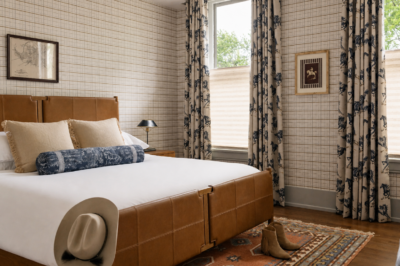 A neatly made bed with brown leather frame, decorative pillows, a hat, and boots beside it; patterned curtains and framed art decorate this inviting Sylvan Park boarding house bedroom.