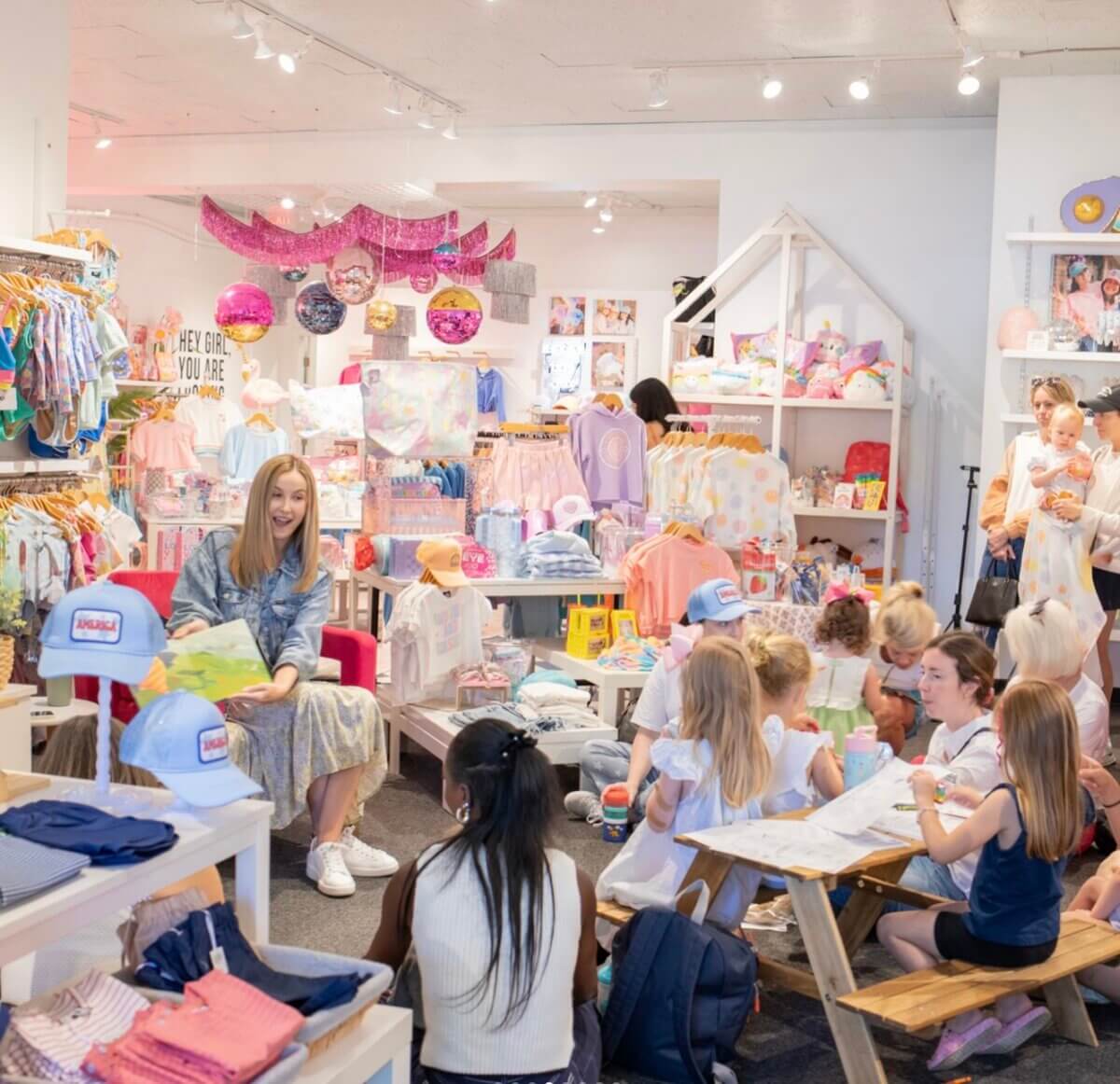 A woman reads a book to a group of children seated on benches in a brightly lit Nashville children's store filled with colorful clothing and toys.