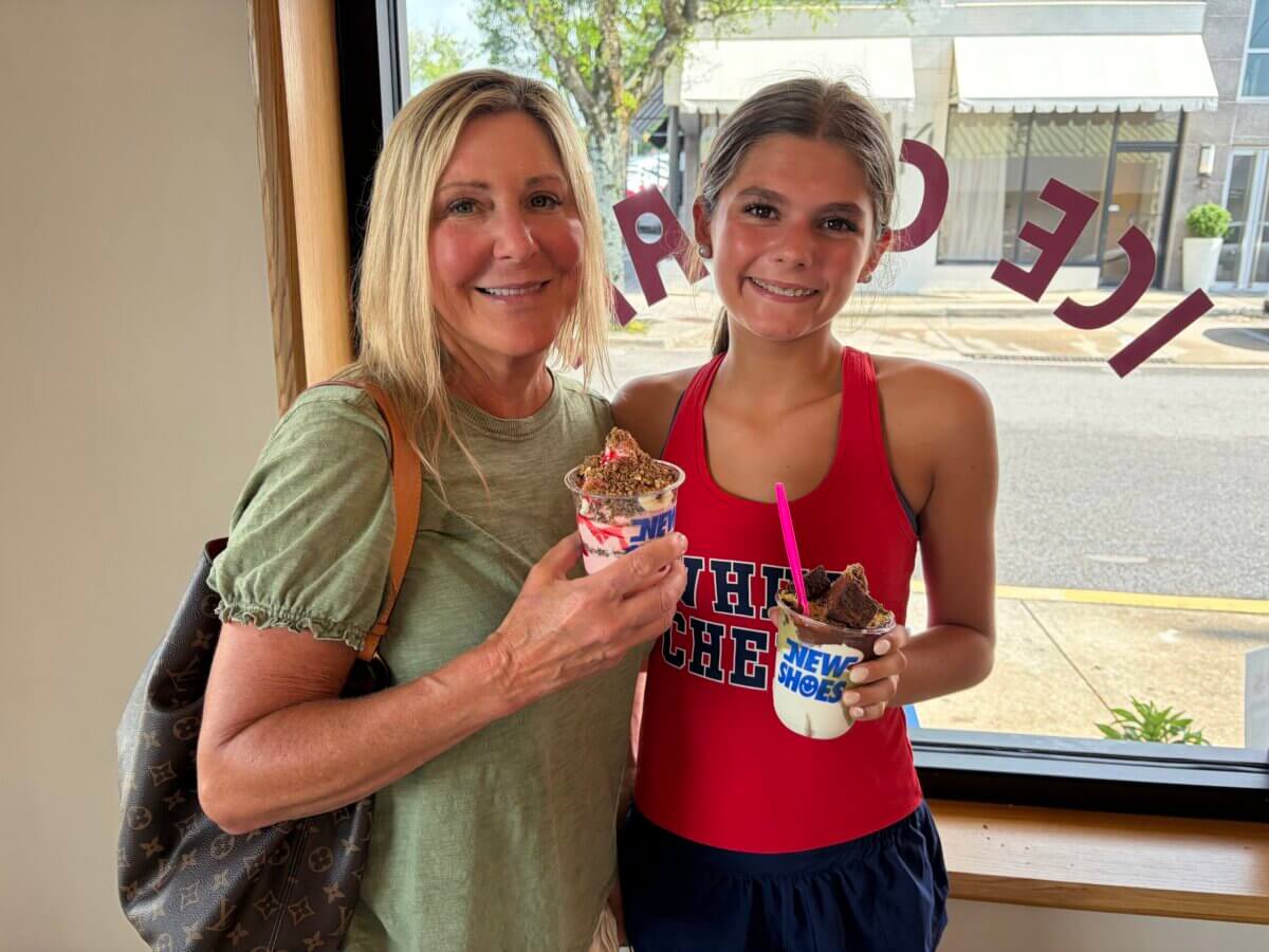 Scoops of Nostalgia: Why Everyone is Lining Up for New Shoes Soft Serve - 4 Two people stand indoors by a window in Homewood, smiling and holding cups of New Shoes Soft Serve with toppings. The window has reverse lettering that reads "ICE CREAM."