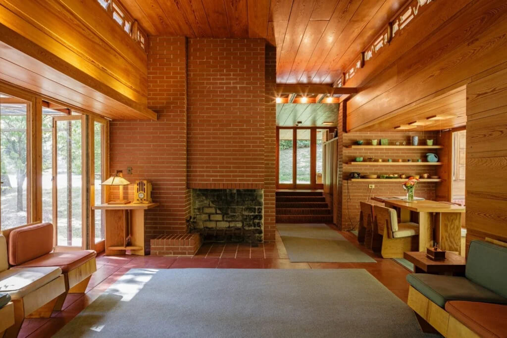 A spacious living room with wood paneling, large brick fireplace, built-in seating, and shelves with pottery, with large windows overlooking trees.