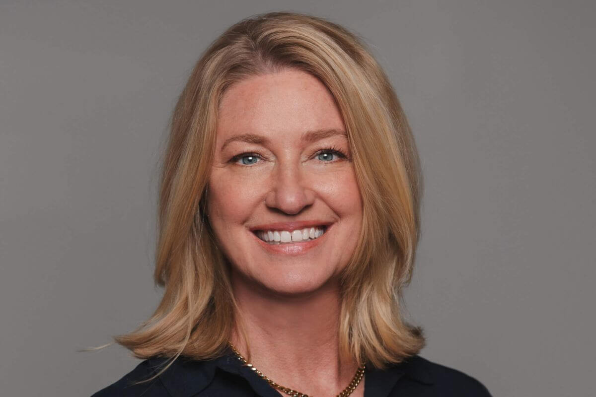 A woman with shoulder-length blonde hair wearing a navy top and gold necklace, smiling at the camera against a plain gray background.