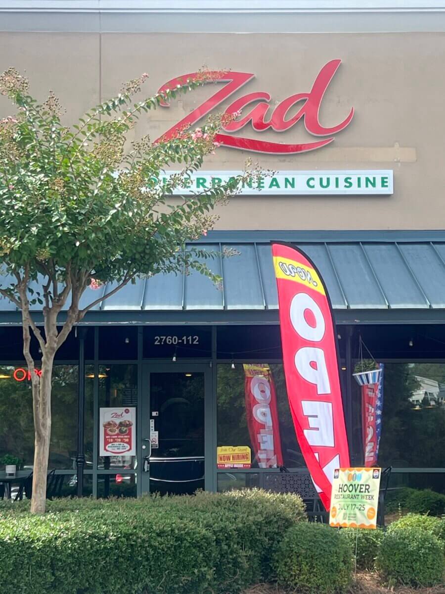 11 New Birmingham Restaurants to Try This Month - 10 Storefront of Zad Mediterranean Cuisine, one of the exciting new restaurants in Birmingham, with a red "Open" flag, a tree on the left, and signs in front of the entrance advertising the restaurant’s offerings and location.