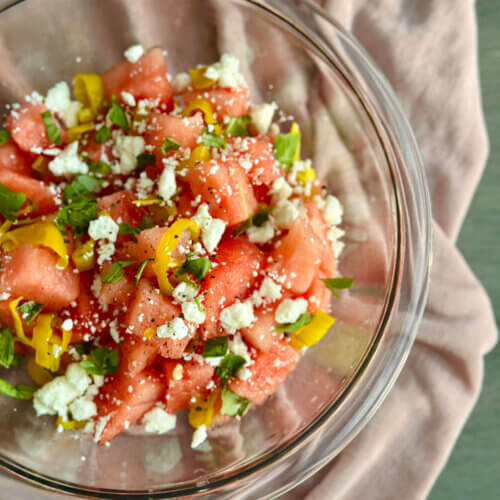 RECIPE: Watermelon Pepperoncini Salad - 3 A glass bowl of watermelon salad topped with crumbled feta cheese, sliced yellow peppers, and fresh herbs, placed on a light pink cloth.