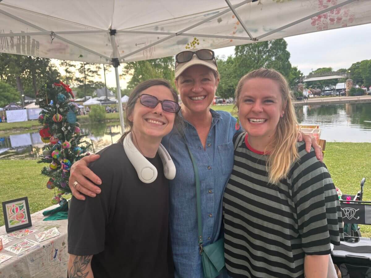 Three smiling women stand together outdoors under a canopy near a decorated table and a pond, with trees and tents visible in the background.