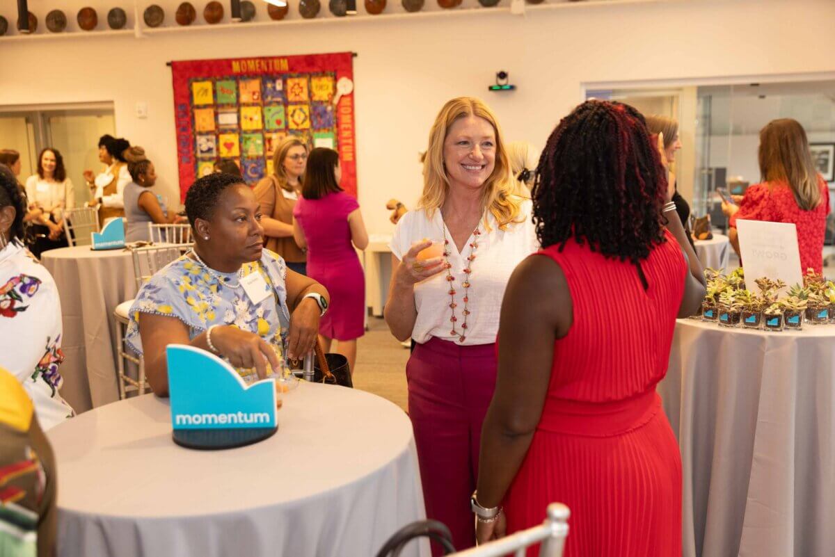 A group of women stand and talk around cocktail tables at a networking event; April Benetollo is among them, and a "momentum" sign is visible on a table.