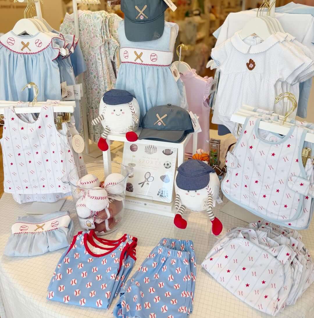 8 Birmingham Children's Boutiques for Every Age - 2 Display of children’s clothing and accessories with baseball and sports motifs, including shirts, shorts, caps, bibs, and plush dolls on a store table at Once Upon a Time.