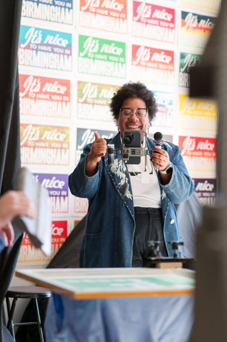 She Connects BHAM Businesses, Brews & Babes - 6 Kayla Stinnett smiles and gives a thumbs up while holding a vintage camera, standing in front of a colorful wall with repeating "It's nice to have you in Birmingham" posters.