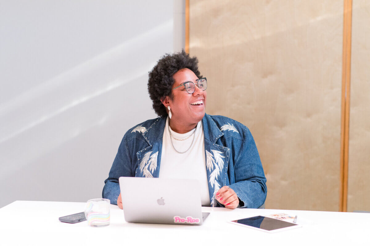 She Connects BHAM Businesses, Brews & Babes - 2 Kayla Stinnett sits at a desk smiling, with a laptop, smartphone, tablet, and a glass of water in front of her at Iron City Social.