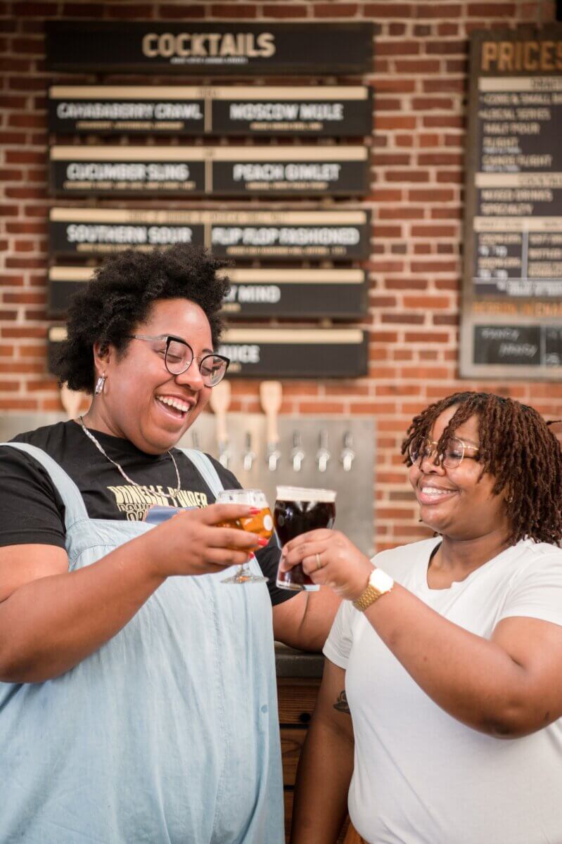 She Connects BHAM Businesses, Brews & Babes - 4 Kayla Stinnett and a friend stand indoors, smiling and clinking their drinks together in front of a bar, with a cocktail menu and drink taps visible in the background.