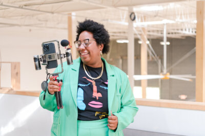 Kayla Stinnett from Iron City Social, dressed in a green suit, holds a smartphone mounted on a handheld tripod with a microphone, smiling indoors. Image by Deborah Michelle Photography.
