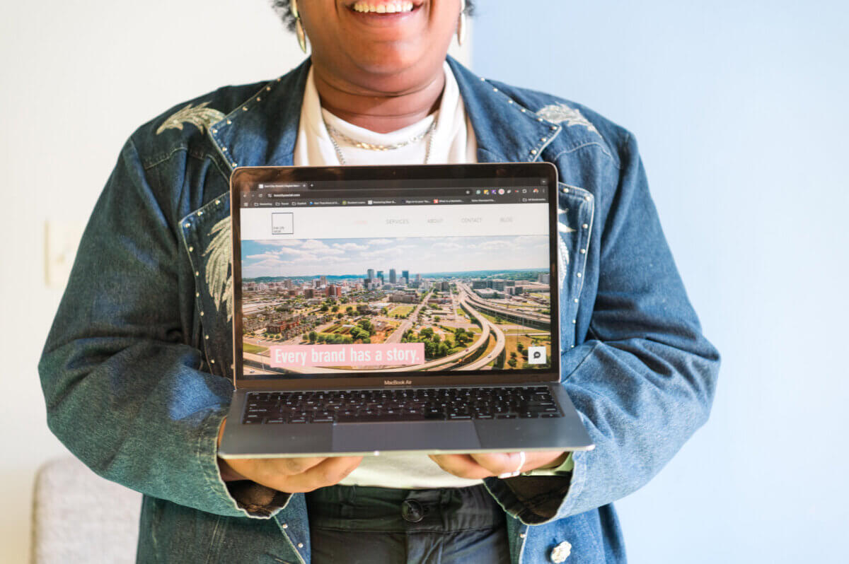 She Connects BHAM Businesses, Brews & Babes - 3 Person holding an open laptop displaying a website with a cityscape and the text "Every brand has a story," created by Iron City Social and Kayla Stinnett.