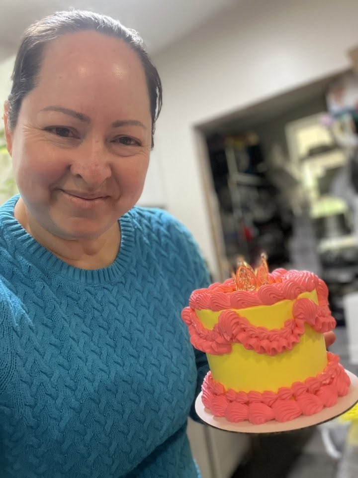 Meet the Internet Star Behind BHAM’s FIRST Mexican Mobile Bakery! - 2 Ana Araujo in a blue sweater holds a small, decorated yellow cake with pink frosting and a candle in a kitchen setting.