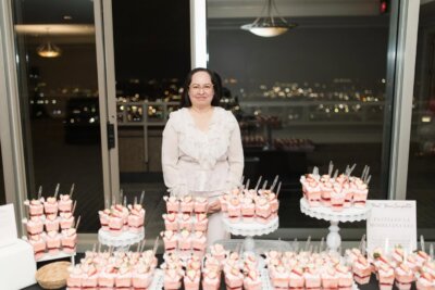 Ana Araujo stands behind a table displaying multiple trays of pink desserts in clear cups, each topped with strawberries and small spoons. Behind her, the BHAM cityscape is visible.