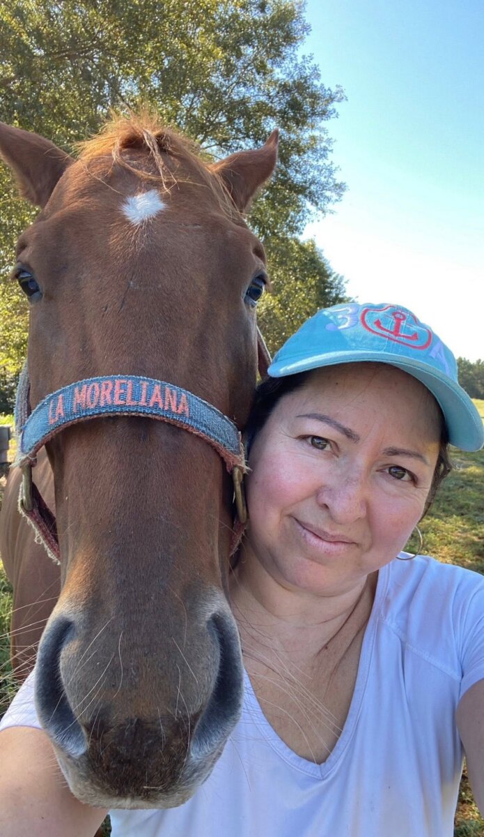 Meet the Internet Star Behind BHAM’s FIRST Mexican Mobile Bakery! - 9 Ana Araujo blue cap takes a close-up selfie with a brown horse wearing a halter labeled "LA MORELIANA," outdoors on a sunny day.
