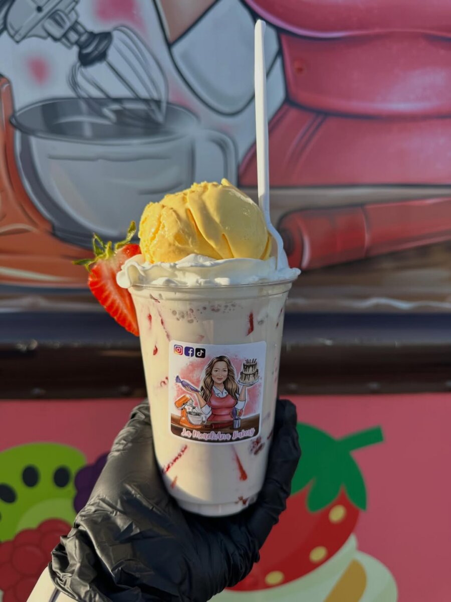 Meet the Internet Star Behind BHAM’s FIRST Mexican Mobile Bakery! - 7 A gloved hand holds a sundae topped with a scoop of yellow ice cream, whipped cream, and a strawberry, against the colorful mural background of La MoreliAna Food Truck, BHAM’s favorite Mexican Mobile Bakery.