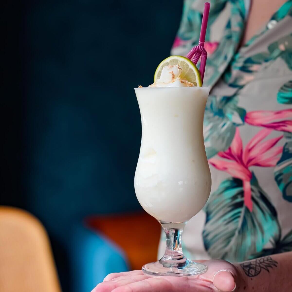 10 Frozen Cocktails to Try in BHAM This Summer - 5 A person in a floral shirt holds a tall glass a pina colada frozen cocktail, garnished with a lime slice and straw from Cayo Coco.