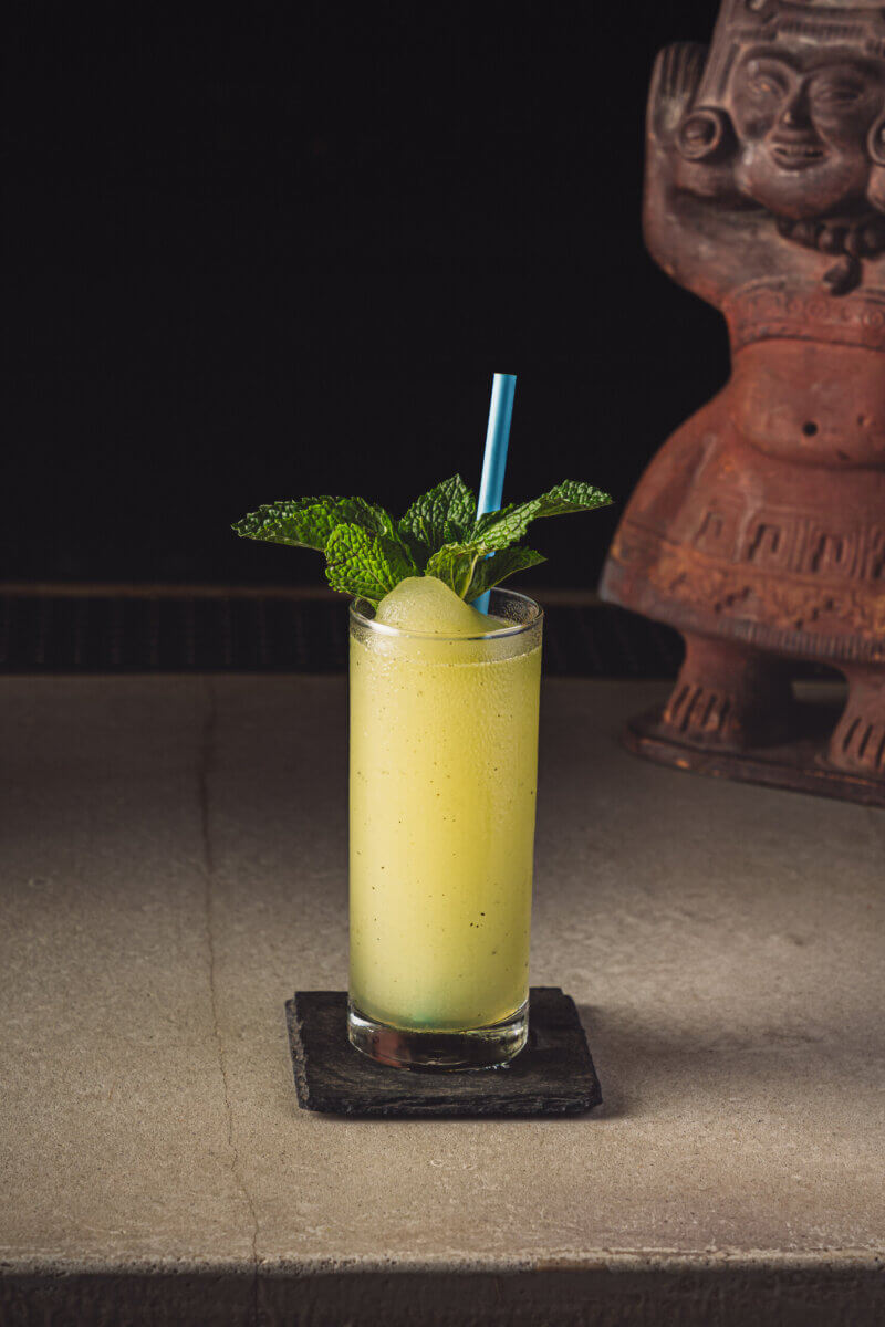 10 Frozen Cocktails to Try in BHAM This Summer - 7 A tall glass of yellow frozen cocktail, garnished with mint leaves and a blue straw, sits on a stone coaster at Adios Bar.