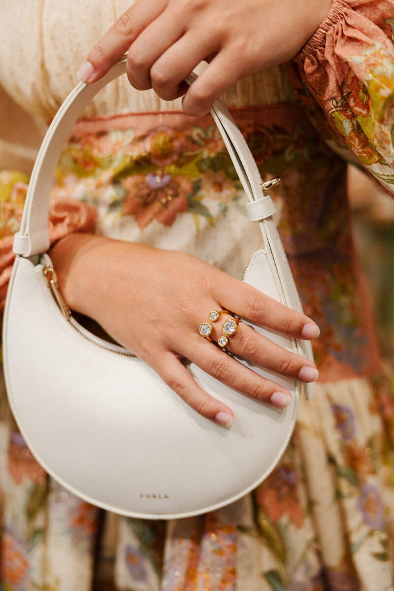 5 Formalwear Trends You'll See Everywhere This Summer - 3 A person wearing a floral dress holds a white Furla handbag, showcasing two gemstone gold rings β an elegant nod to Birmingham formalwear trends.