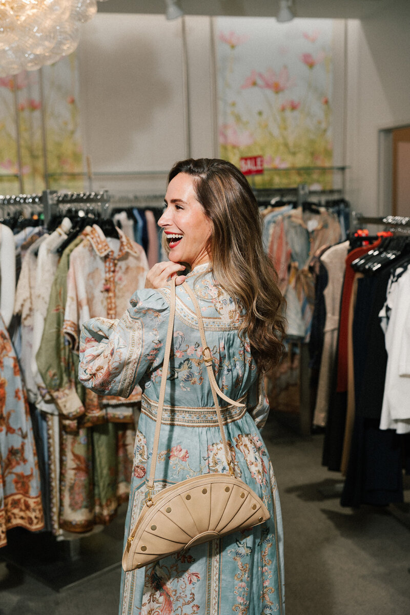 5 Formalwear Trends You'll See Everywhere This Summer - 4 A woman wearing a patterned dress and holding a beige purse over her shoulder stands in Gus Mayer, a boutique clothing store in Birmingham, smiling and looking back at the latest formalwear trends.