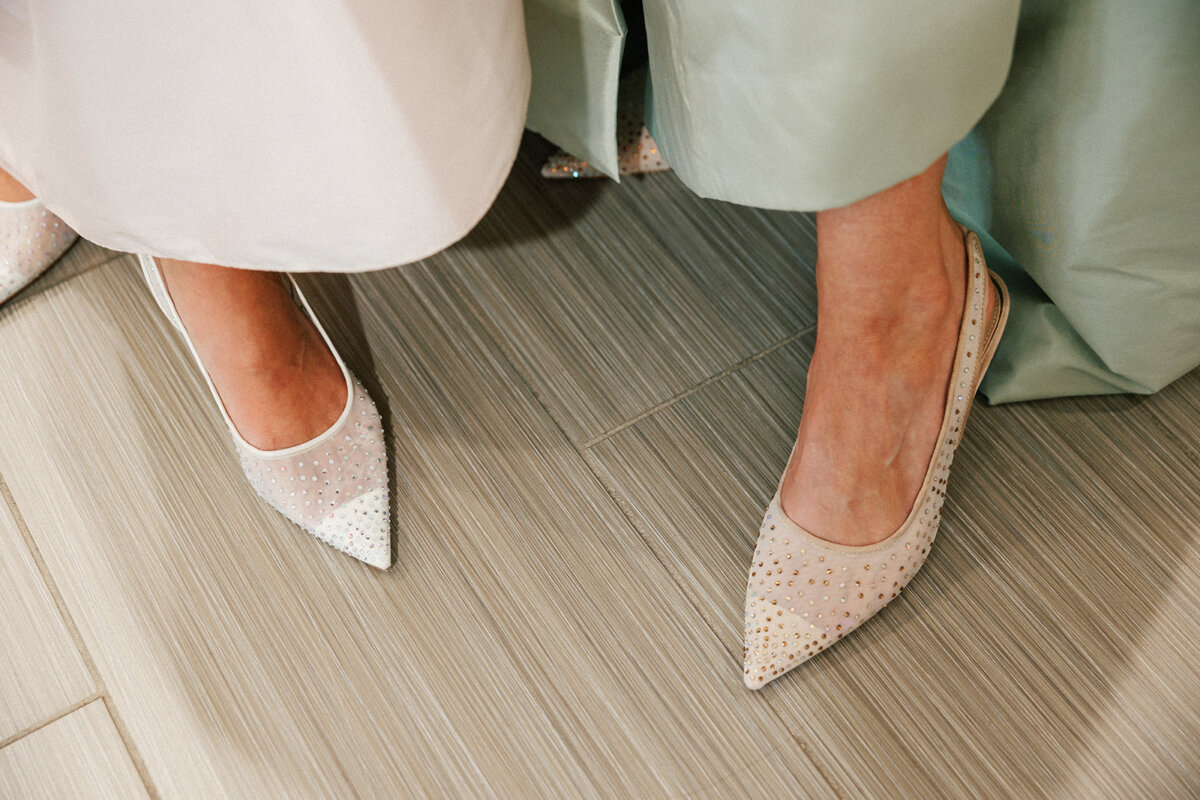 5 Formalwear Trends You'll See Everywhere This Summer - 7 Two people wearing light-colored, pointed-toe shoes decorated with small rhinestones β reflecting the latest Formalwear Trends β stand on a light wood-patterned floor.