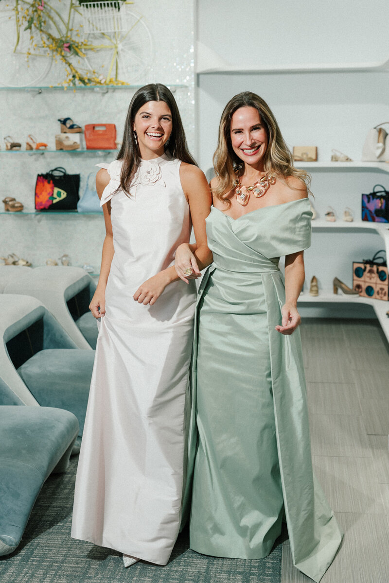 5 Formalwear Trends You'll See Everywhere This Summer - 5 Two women stand smiling indoors in elegant floor-length gowns β one in white, the other in light green β showcasing Birmingham Formalwear trends. Shelves with shoes and handbags are visible in the background.