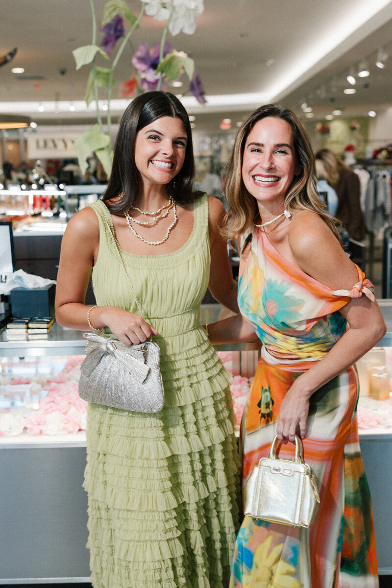 5 Formalwear Trends You'll See Everywhere This Summer - 8 Two women smile together indoors, showcasing Birmingham Formalwear. One wears a light green ruffled dress with a silver bag from Gus Mayer, while the other stuns in a colorful floral dress and gold purse, reflecting the latest formalwear trends.
